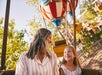 A woman and a young girl smile at each other while sitting on a carnival ride with colorful gondolas. Trees and a clear sky are in the background.