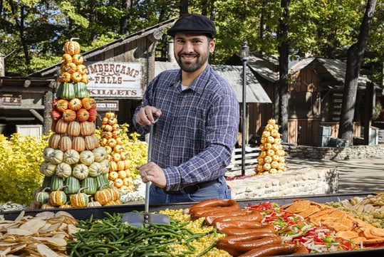 A man in a plaid shirt and flat cap cooks sausages, peppers, and other vegetables outdoors at a food stand, with stacked gourds and rustic buildings in the background.