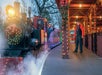 A festive train adorned with holiday lights emits steam. One person stands by the engine, while another stands on the platform, both dressed in uniform.