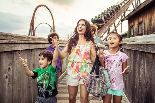 A woman and three children walk on a wooden walkway near a roller coaster, looking excited and pointing at something off-camera.