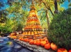 A large, tiered pyramid made of stacked pumpkins and gourds stands outdoors, surrounded by trees, bushes, and more pumpkins on the ground.