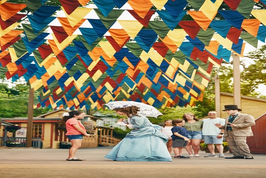 A woman in a Victorian-style dress and man in period attire greet a family under colorful triangular banners outdoors.