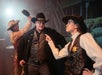 Three people in Western costumes: a sheriff points at a man in black, while another person raises a bottle behind the man's head as if about to strike.