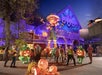 Halloween-themed scene at a theme park with a large pumpkin figure, glowing jack-o'-lanterns, and people in colorful costumes under a clear night sky.