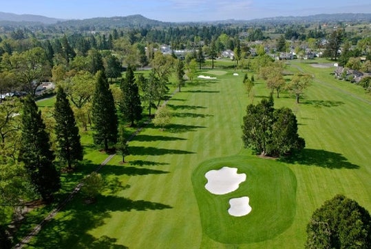 Aerial view of a golf course with green fairways, several sand bunkers, and surrounding trees on a sunny day. Houses and hills are visible in the background.