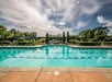 Outdoor swimming pool with clear blue water, surrounded by greenery and a landscaped patio, under a partly cloudy sky.