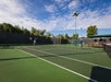 Two people playing tennis on an outdoor court. The sky is clear with some clouds, and trees are visible in the background.