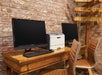 Two computer monitors with keyboards and a printer on a wooden desk against a brick wall. Two brown chairs are in front of the desk. A directional wooden signpost is in the background.