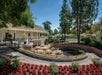A landscaped garden with a white building, red flowers, and a set of round tables set on a patio. Tall trees and a tennis court are visible in the background under a clear blue sky.
