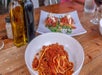 A bowl of pasta with meat sauce is in the foreground. In the background, a plate of bruschetta is visible alongside bottles of olive oil and vinegar on a wooden table.