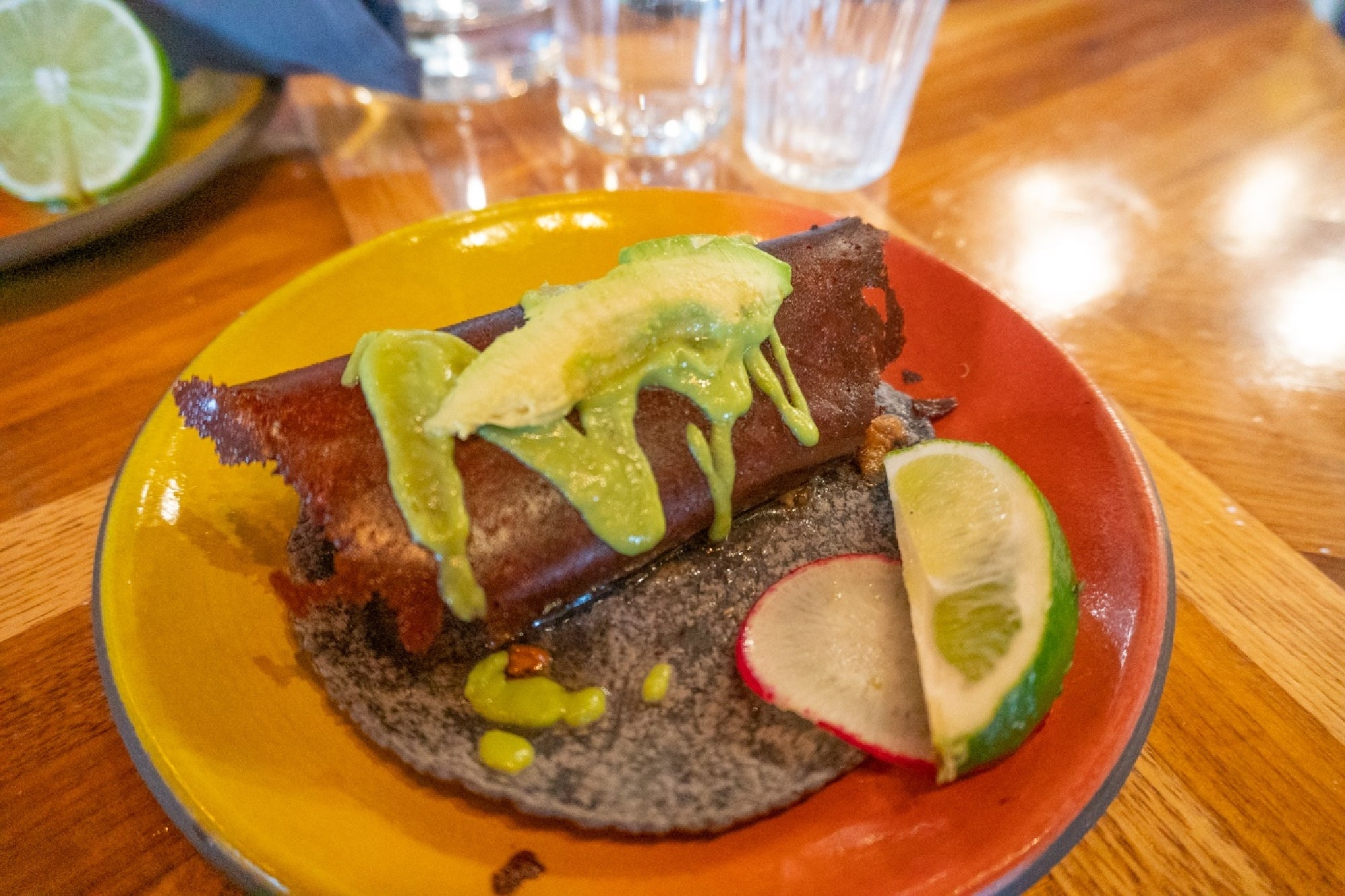 A plated taco with a crispy shell, topped with avocado and green sauce, served with a slice of lime and a radish on a multicolored plate.