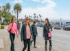 People walk on a sunny sidewalk by a beach road lined with palm trees, cars parked nearby.