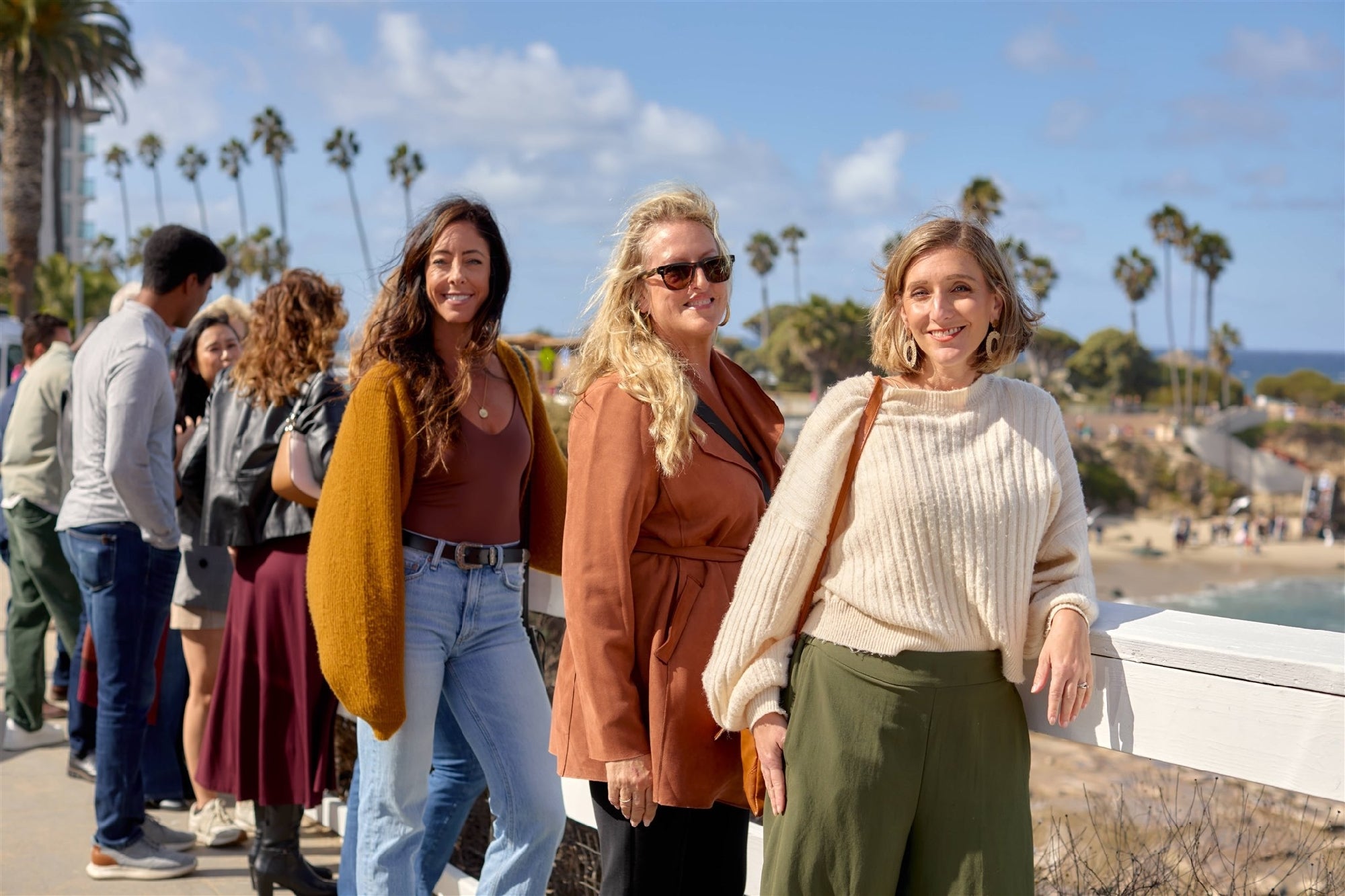 Three women stand by a white railing at a beachside viewpoint on a sunny day, with other people and palm trees in the background.