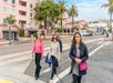 A group of four women with long hair walking from the crosswalk to the sidewalk with a pink building and palm trees behind them.
