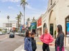 A group of women standing on the sidewalk and listening to their tour guide with stores in the background on a sunny day in San Diego.