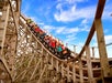 A group of people ride a wooden roller coaster with their hands up as the train descends a steep curve under a blue sky with scattered clouds.