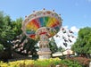 People ride a colorful swing carousel in an amusement park surrounded by greenery and flowers under a blue sky.