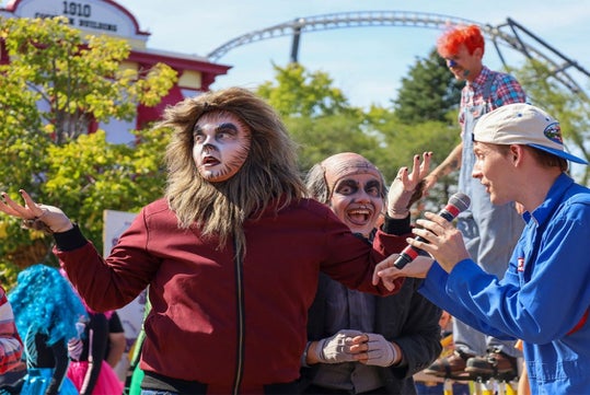 People in costumes, including a person dressed as a lion, perform outdoors. A person with a microphone speaks, while a roller coaster is visible in the background.