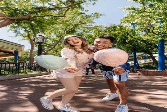 A couple smiles for picture while holding a pink cotton candy inside Six Flags America with trees behind them.