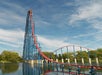 Roller coaster with a steep blue and red track, ascending high over a body of water, set against a partly cloudy sky and surrounded by trees.