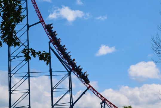 People riding a steep roller coaster on a sunny day with scattered clouds, surrounded by some tree branches.