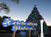 A tall Christmas tree decorated with colorful lights stands behind a blue sign that reads "Celebration Station." A person is standing nearby in the foreground.
