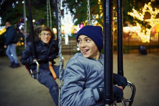 Two boys wearing winter jackets and hats ride on swings at an outdoor amusement park at night, with festive lights and blurred people in the background.