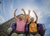 Two people on a roller coaster raise their arms and smile, with a blue sky and wooden structure in the background.