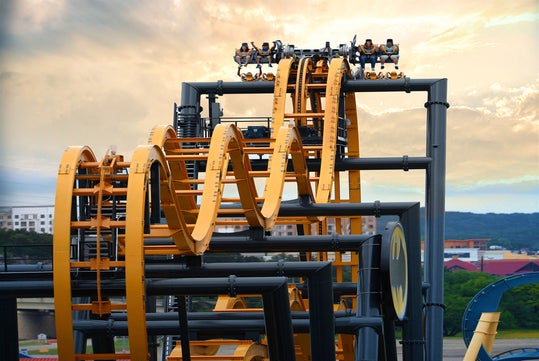 A roller coaster with an intricate loop track carries riders, set against a cloudy sky and distant urban background.