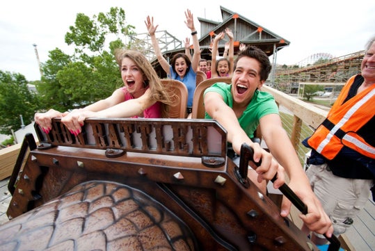 A group of people ride a wooden roller coaster with excited expressions, gripping the safety bar; a ride operator stands nearby in a safety vest.