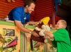 A carnival worker hands a young boy a stuffed monkey prize at an arcade game booth. Both are smiling and the booth displays colorful decorations and scoreboards.