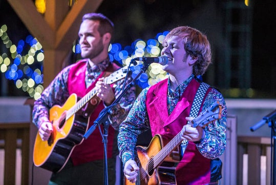Two musicians wearing red vests and patterned shirts play acoustic guitars and sing into microphones on an outdoor stage with string lights in the background.