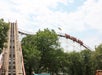 A wooden roller coaster with a steep incline on the left. Riders are visible on the right track section, surrounded by green trees under a partly cloudy sky.