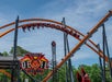 People ride the Jersey Devil Coaster, an orange steel roller coaster with a devil-themed sign, under a blue sky with trees in the background.