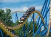 People ride a large yellow and blue roller coaster as it goes over a curved track outdoors on a sunny day, with trees and blue sky in the background.