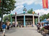 A colorful carousel with striped pillars and a pointed roof stands in an amusement park, with people walking nearby and trees in the background.