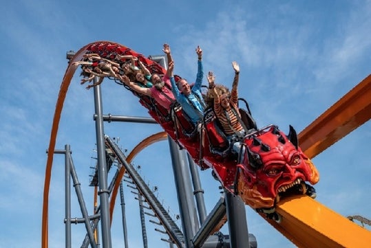 Roller coaster with a red devil-themed front car, carrying passengers with raised arms, ascending a steep track against a clear blue sky.