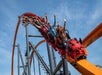 Roller coaster with a red devil-themed front car, carrying passengers with raised arms, ascending a steep track against a clear blue sky.