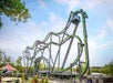 Tall, green and purple looping roller coaster with multiple twists and turns under a blue sky. Nearby, a striped tent and trees are visible.