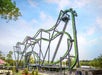 Large steel roller coaster with green and purple tracks featuring steep drops, loops, and twists, set outdoors in an amusement park under a partly cloudy sky.