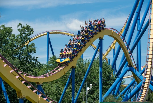 A roller coaster with a yellow track and blue supports carries riders over a loop in a theme park, with some people raising their hands. Trees and a blue sky are in the background.