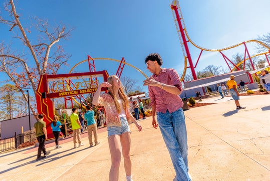 Two people walk and talk in front of a roller coaster called "Vertical Velocity" at an amusement park on a sunny day. Other visitors are gathered near the ride entrance.