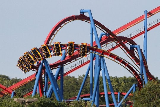 A roller coaster with multiple loops and blue support beams is pictured against a clear sky, with trees visible at the bottom.