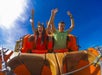 Two people ride a roller coaster, arms raised and smiling, against a clear blue sky.