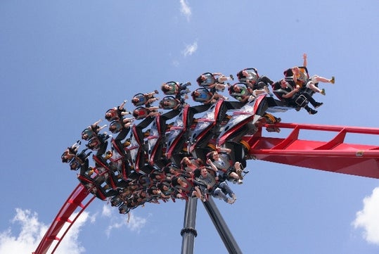 Roller coaster sharply turns upside down as riders, secured in seats, raise their hands and scream against a background of blue sky and clouds.