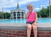 A young child in a pink shirt sits on a brick ledge in front of a large fountain and a carousel, with trees and a blue sky in the background.