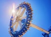A large circular amusement park ride spins in the air under a bright blue sky, with riders’ legs extending outward and sunlight shining through the structure.