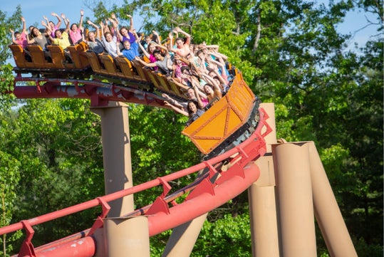A group of people ride a red roller coaster, raising their arms as the train speeds around a curved track surrounded by green trees.