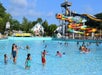 People of various ages swim and play in a large outdoor water park pool with colorful water slides and buildings in the background.
