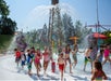 Children run and play under water spraying from a fountain at an outdoor splash pad on a sunny day, surrounded by playground equipment and trees.
